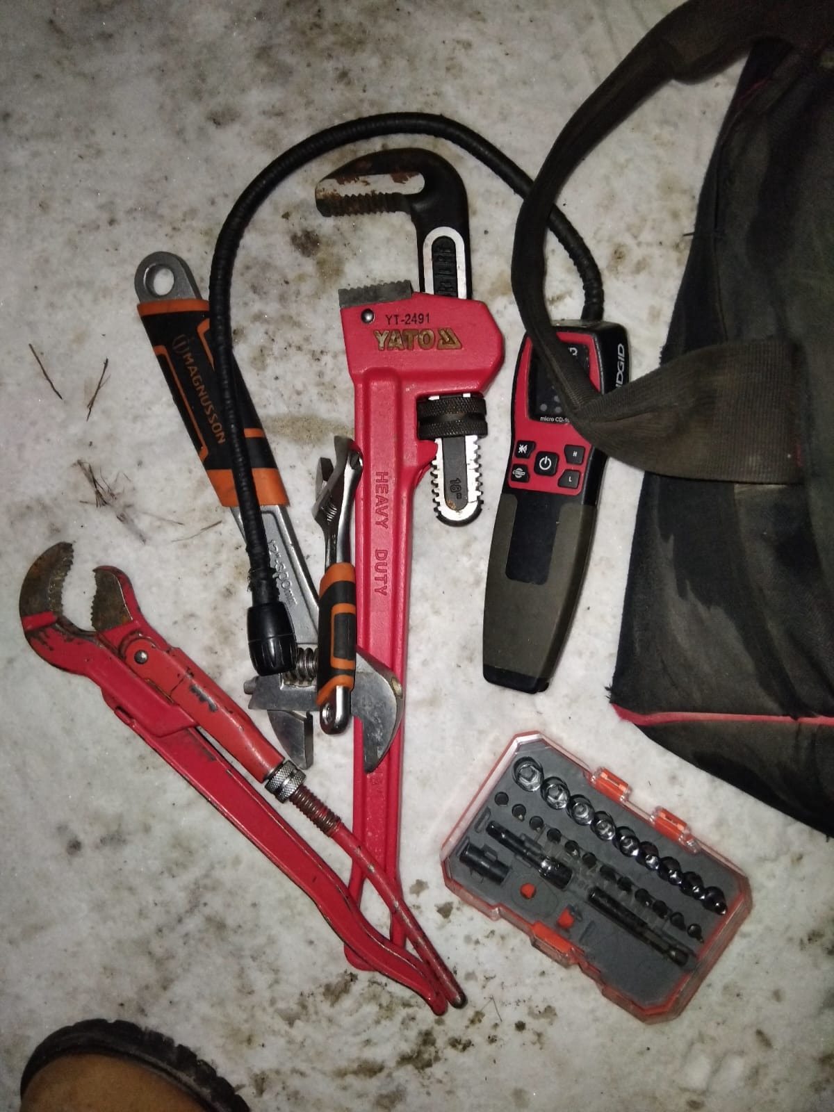 Różnorodne narzędzia gazownika: klucze nastawne, szczypce, zestaw bitów i detektor gazu leżące na podłożu. Widoczny fragment torby narzędziowej.