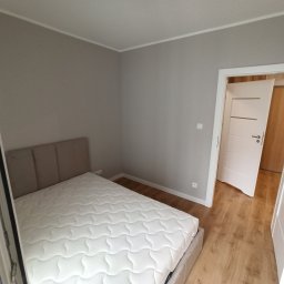 Michał Wiśniewski - Jasne wnętrze sypialni po remoncie: beżowe łóżko z materacem, drewniana podłoga i białe drzwi z poziomymi frezami. Ściany pomalowane na szaro.