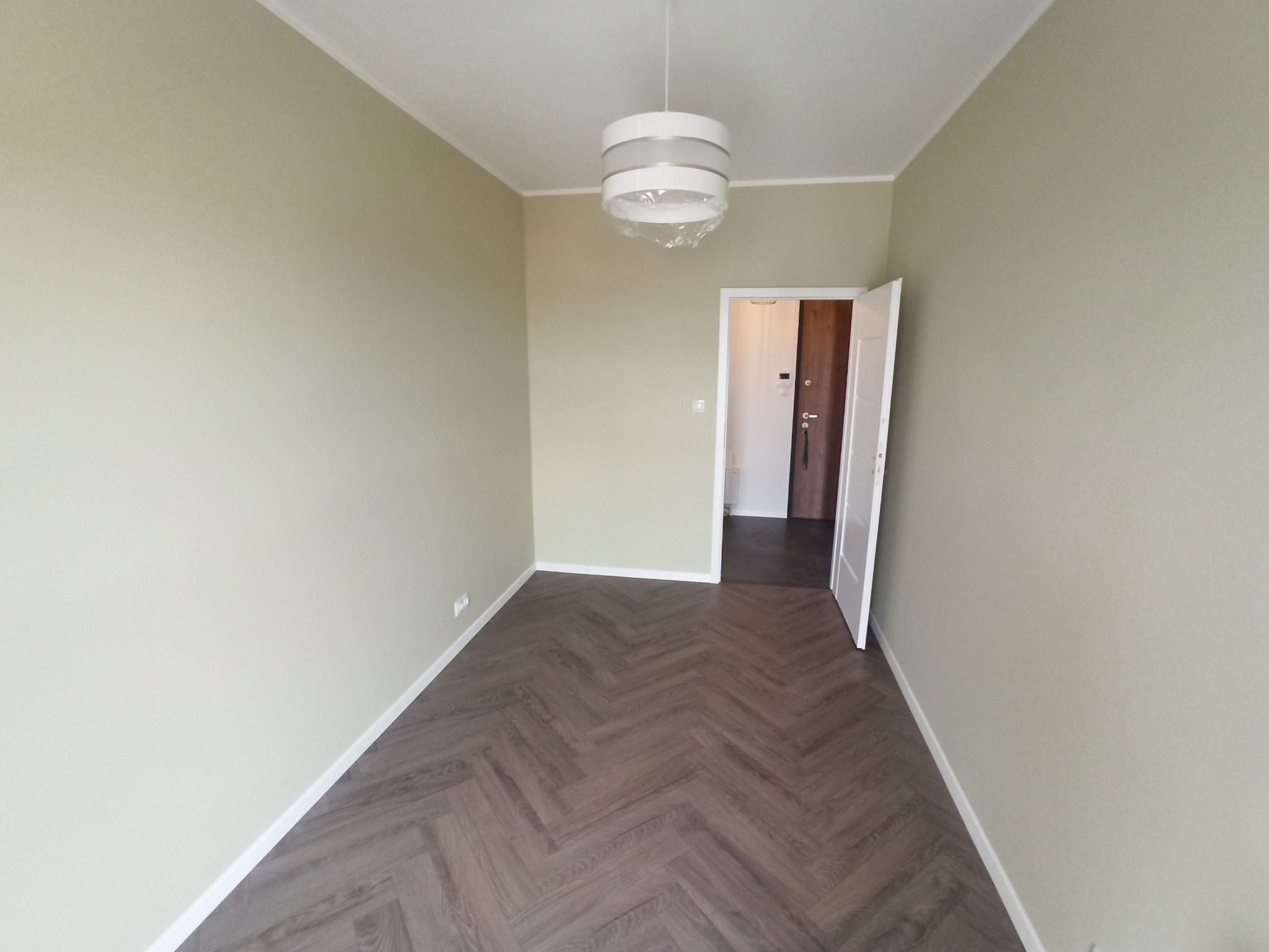 Pomalowane na beżowo puste pomieszczenie z parkietem w jodełkę, z widoczną lampą w folii i otwartymi drzwiami wejściowymi.