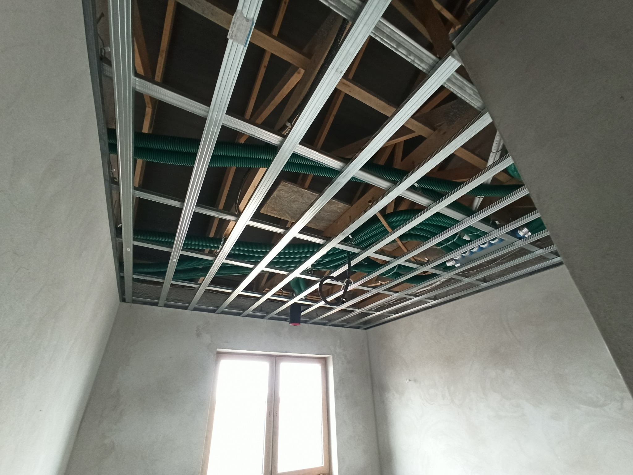 Sufit podwieszany z metalową konstrukcją i zielonymi rurami wentylacyjnymi, widok z dołu, surowe ściany i okno w tle. Etap prac wykończeniowych.
