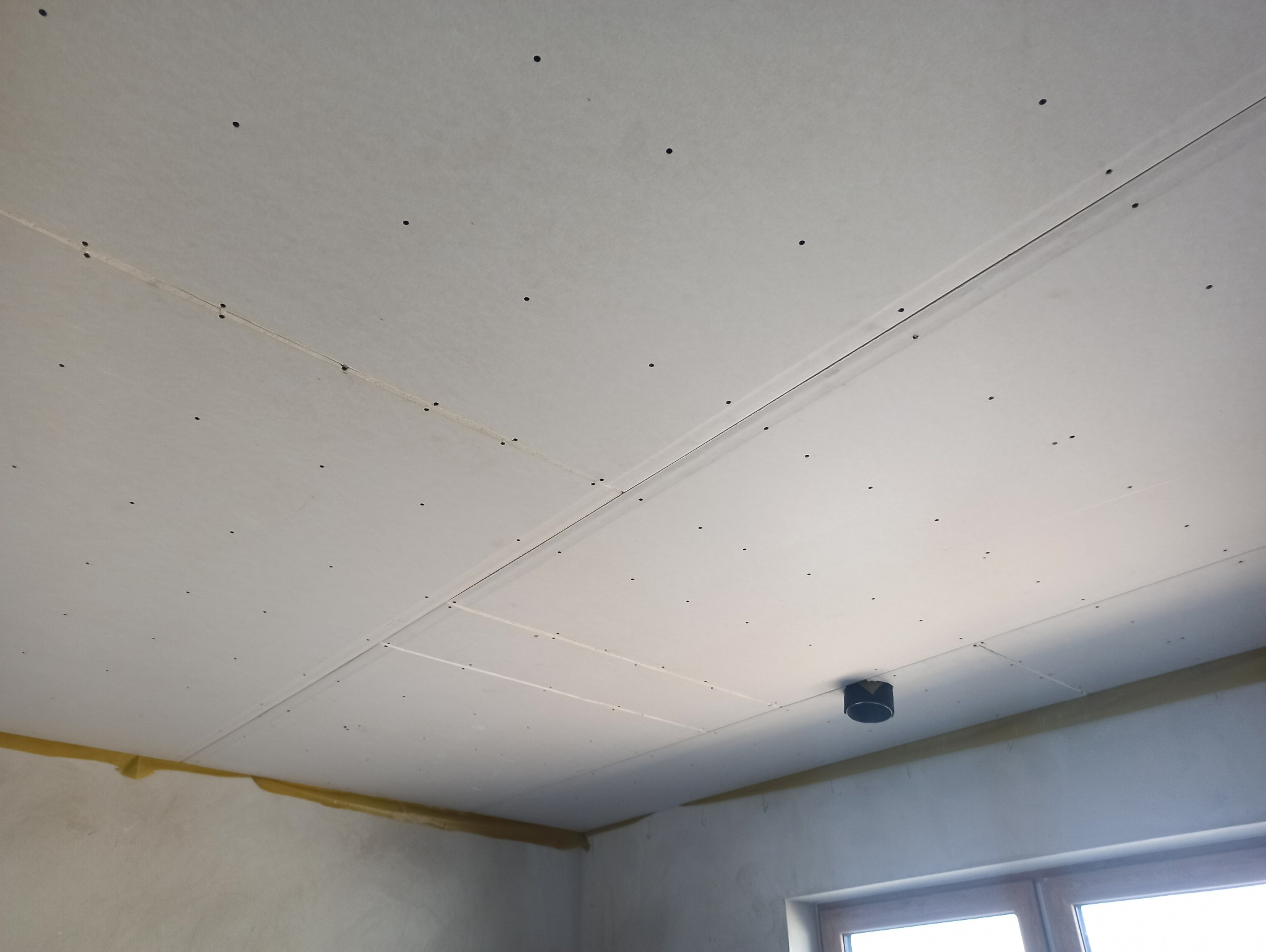 Sufit z płyt kartonowo-gipsowych, widoczne łączenia i wkręty, żółta taśma na styku ze ścianą, fragment okna w rogu. Ujęcie z dołu, jasne oświetlenie.