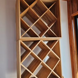 S.P. PALETTEN - LOGISTIC SPÓŁKA Z OGRANICZONĄ ODPOWIEDZIALNOŚCIĄ - Drewniany regał z kwadratowymi przegródkami, ustawiony na tle białej ściany i drewnianej boazerii. Widoczne naturalne usłojenie drewna i precyzyjne wykonanie.