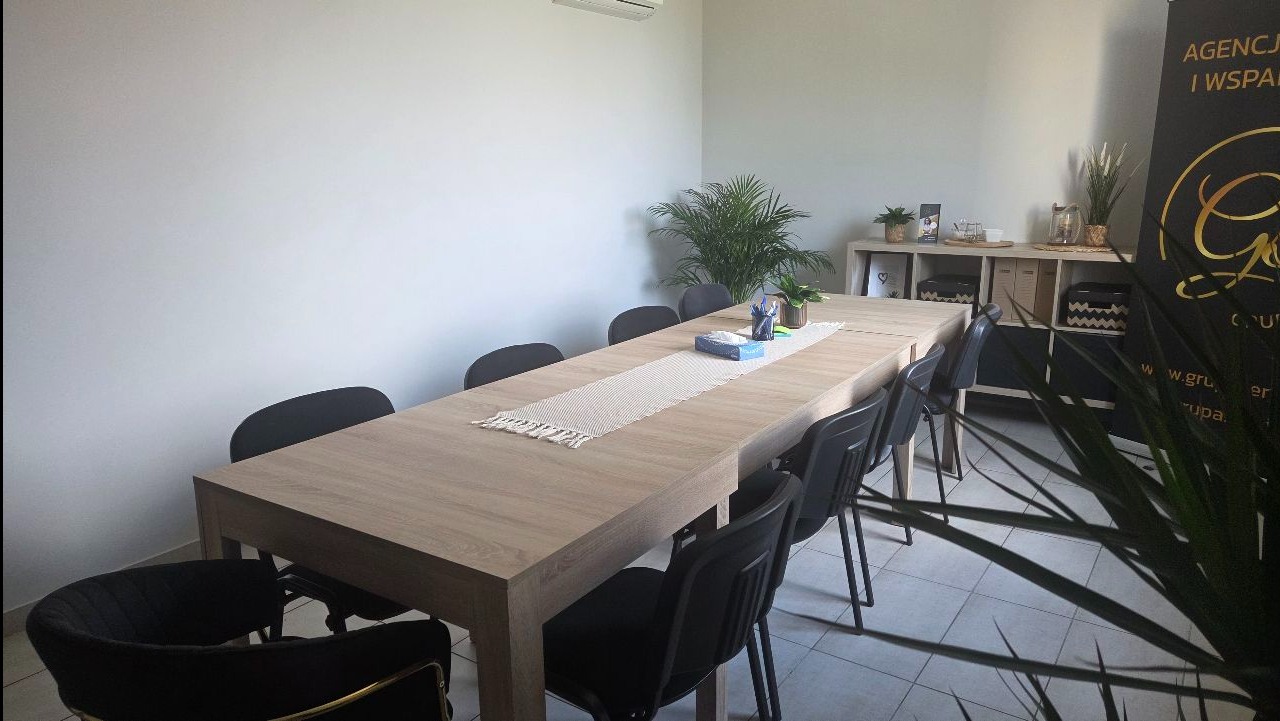 Jasne biuro po sprzątaniu: duży stół konferencyjny z dekoracjami, krzesła, regał z ozdobami, roślina doniczkowa. Czysta przestrzeń gotowa do pracy.