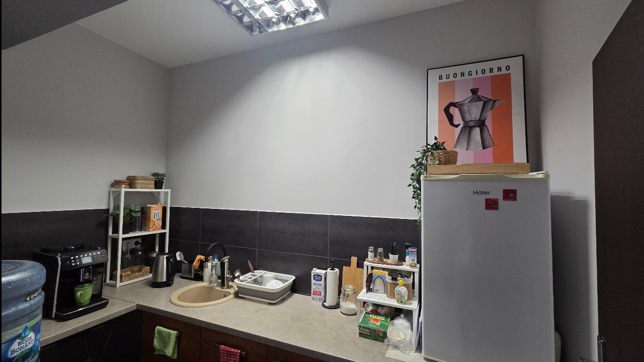 Umyta kuchnia biurowa z ekspresem do kawy, lodówką Haier i plakatem z kawiarką. Czysty blat, umyte naczynia w suszarce. Świeżość i porządek w miejscu pracy.