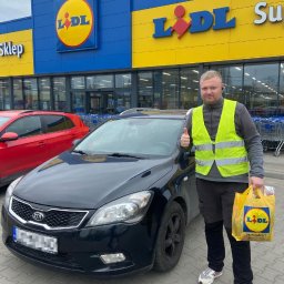 WIKAM - Mężczyzna w kamizelce odblaskowej z torbą zakupową stoi obok czarnego samochodu na tle sklepu Lidl, pokazując kciuk w górę.