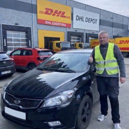 WIKAM - Dostawca w kamizelce odblaskowej, z uniesionym kciukiem, stoi obok czarnego samochodu na tle magazynu DHL. Widoczne logo DHL i inne pojazdy.