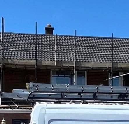 Dom z ciemną dachówką w trakcie prac dekarskich, widoczne rusztowania i drabina na dachu białego samochodu. Błękitne niebo w tle.