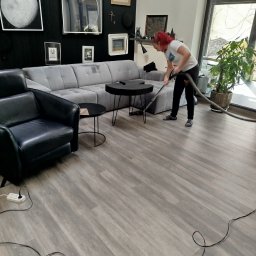 Patrycja Simińska - Kobieta z czerwonymi włosami odkurza podłogę w salonie z szarą sofą i czarnym fotelem. Na ścianie wiszą obrazy, a obok okna stoi roślina doniczkowa.
