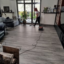 Patrycja Simińska - Kobieta odkurza podłogę w przestronnym salonie z szarymi panelami, używając odkurzacza z długim kablem. W tle widoczne meble, okno balkonowe i dekoracje.