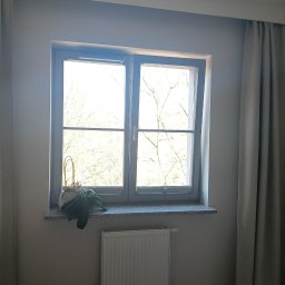 Patrycja Simińska - Wnętrze pokoju po umyciu okna: czysta szyba z widokiem na drzewa, parapet z rośliną, grzejnik i zasłony. Widoczne efekty sprzątania.