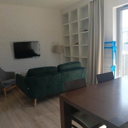 Patrycja Simińska - Jasne wnętrze po sprzątaniu: zielona sofa, szary fotel, stół z krzesłami, biała ściana z cegły i regał. Suszarka na pranie przy oknie balkonowym.