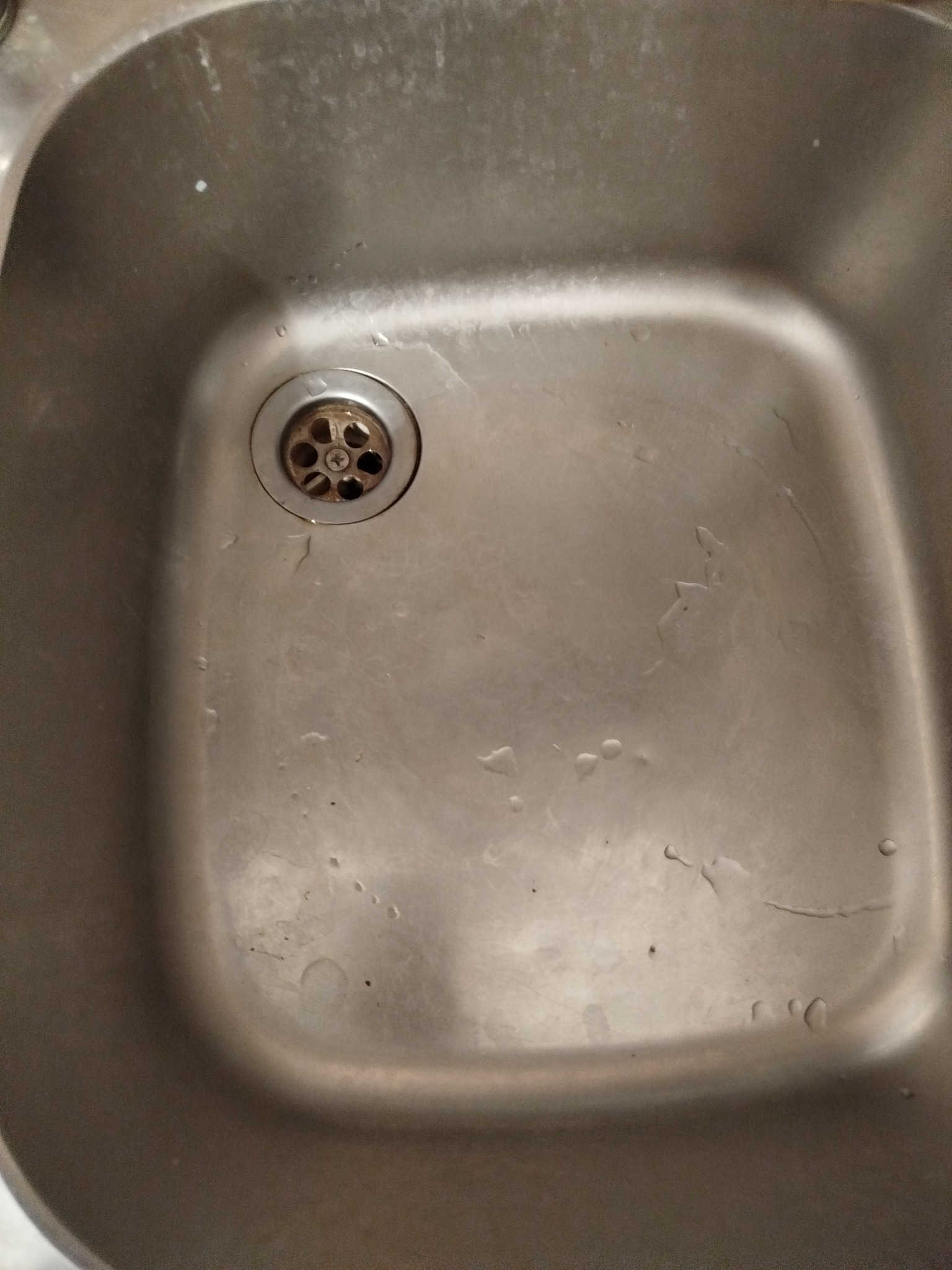 Metalowy zlew kuchenny z widocznymi śladami po wodzie i zabrudzeniami, ukazujący potrzebę czyszczenia. Otwór odpływowy z widocznymi osadami.