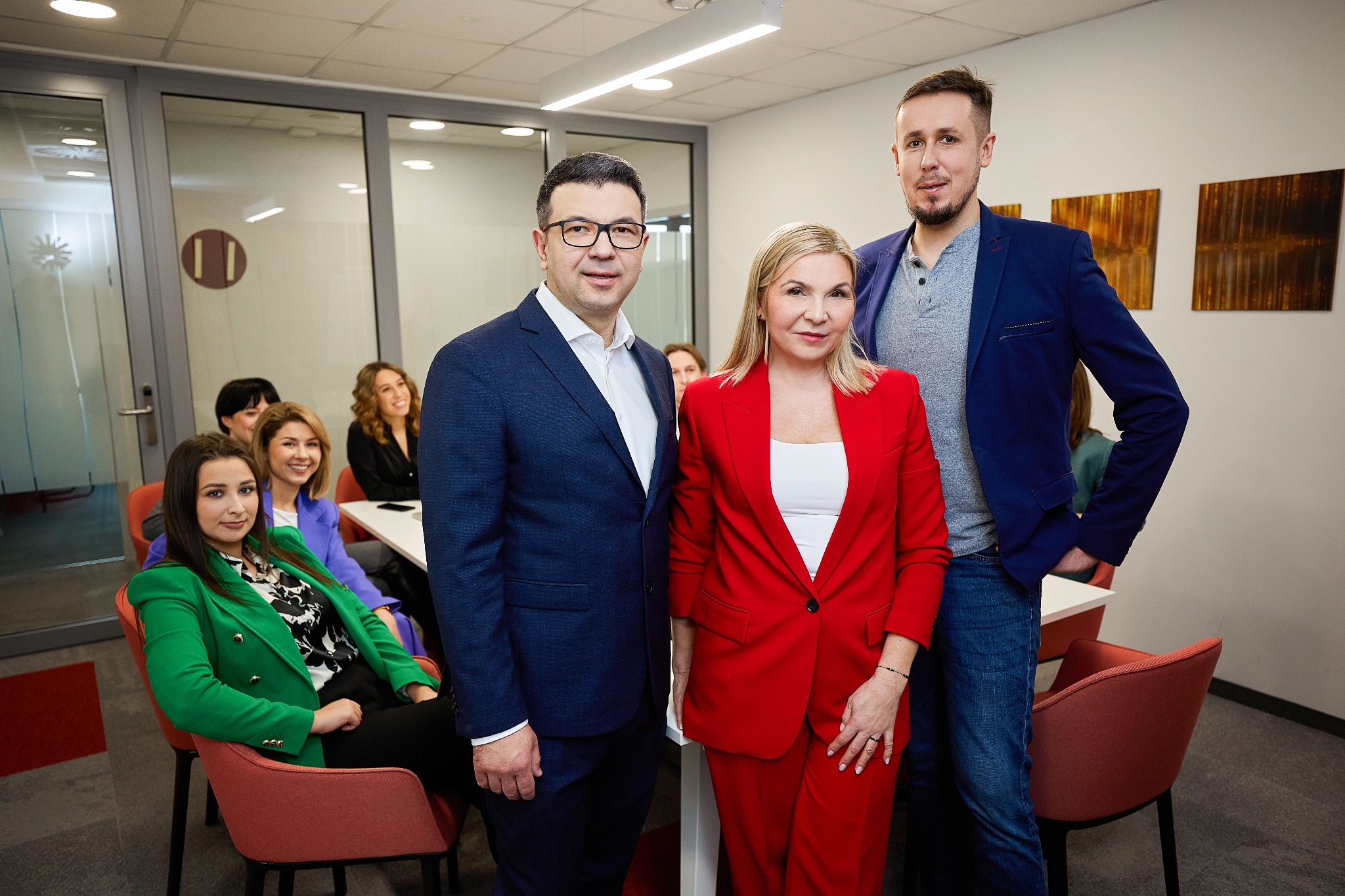 Zespół firmy konsultingowej w biurze: kobieta w czerwonym garniturze, mężczyzna w granatowej marynarce i mężczyzna w niebieskiej marynarce, w tle zespół podczas spotkania.