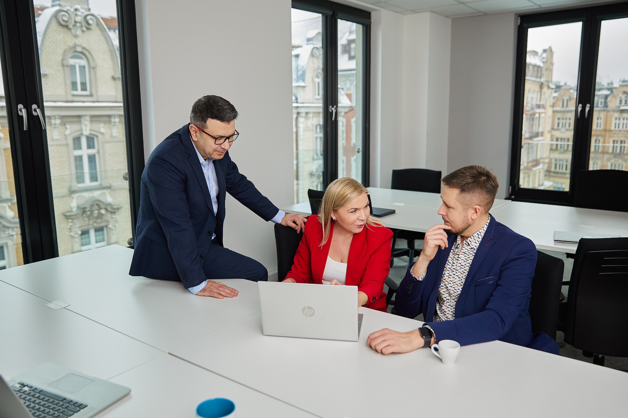 Trzy osoby w biurze omawiające projekt przy laptopie. Mężczyzna oparty o stół, kobieta w czerwonej marynarce i mężczyzna w niebieskiej marynarce. Za oknem widok na miasto.