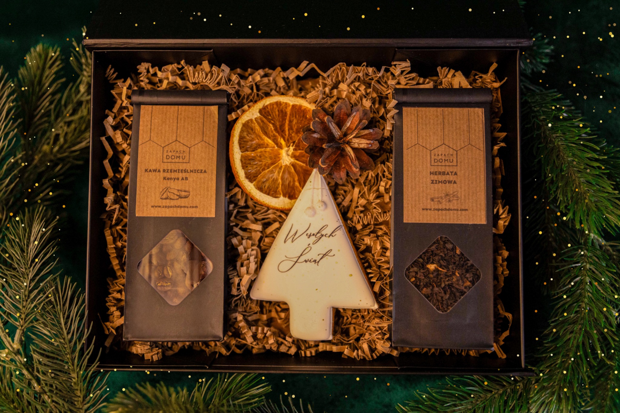 Świąteczny kosz prezentowy z kawą, herbatą, suszoną pomarańczą i piernikiem w kształcie choinki, zapakowany w czarne pudełko z wypełniaczem.