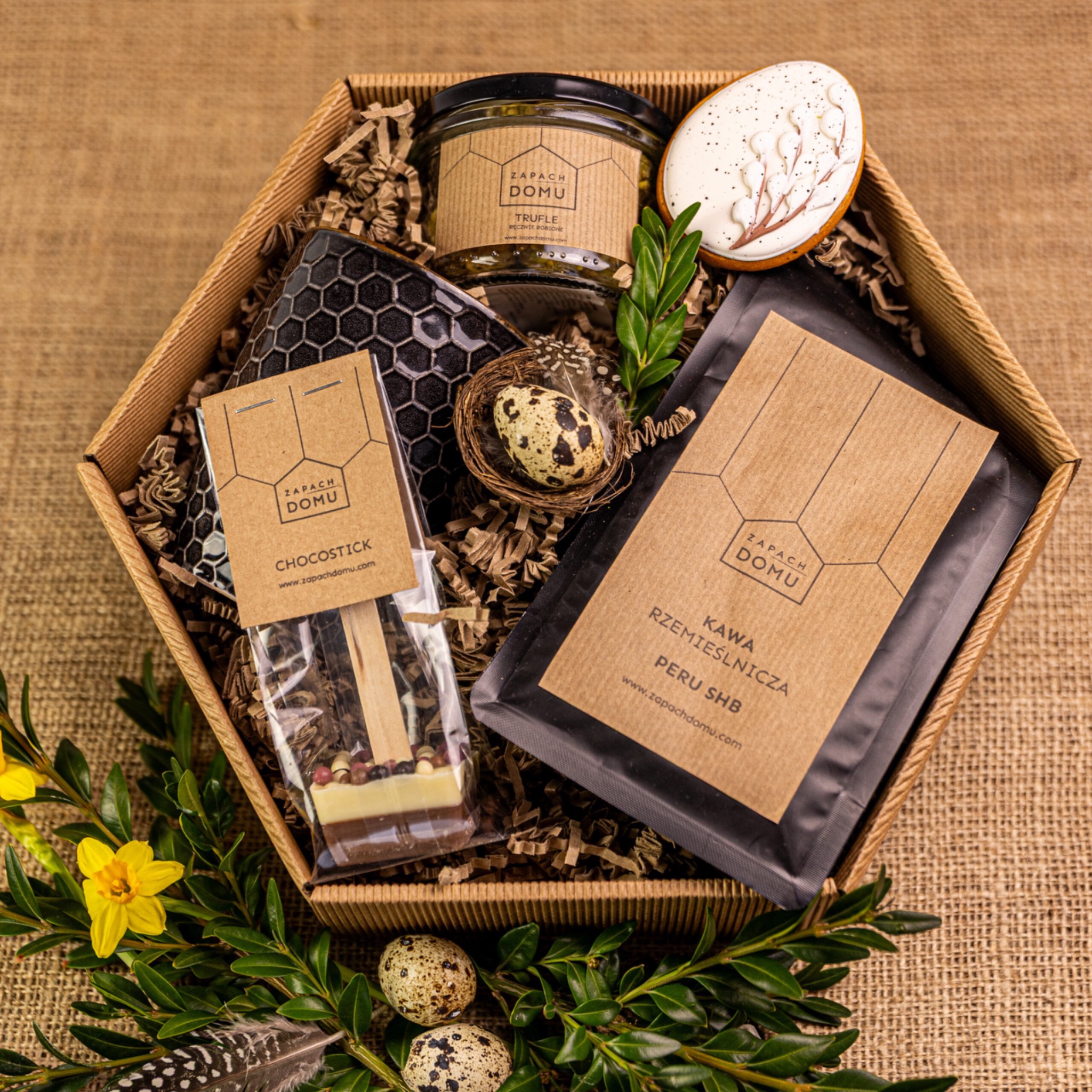 Ozdobny kosz prezentowy z kawą, truflami, czekoladą i dekoracjami wielkanocnymi na jutowym tle, z logo 'Zapach Domu' na produktach.