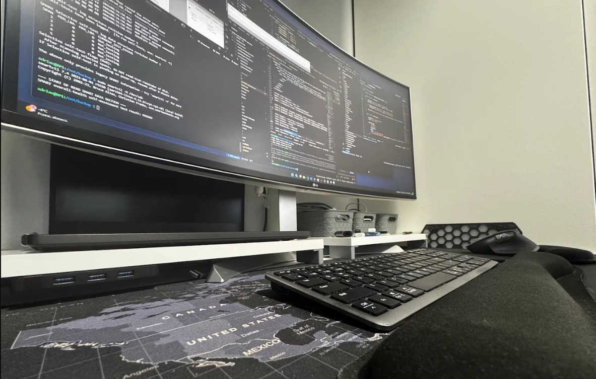 Szeroki monitor LG wyświetla kod, klawiatura i mysz na podkładce z mapą świata. Widok z perspektywy osoby siedzącej przy biurku. Minimalistyczne akcesoria.