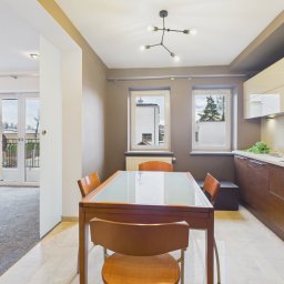 Patrycja Rozpondek NIERUCHOMOŚCI - Jasna jadalnia z drewnianym stołem z szklanym blatem i krzesłami, widok na kuchnię z brązowymi szafkami i salon z balkonem. Nowoczesne oświetlenie sufitowe.