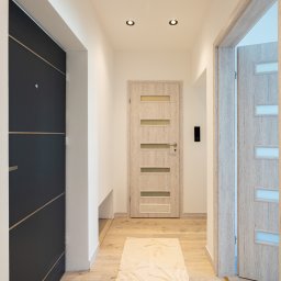 Patrycja Rozpondek NIERUCHOMOŚCI - Nowoczesny korytarz z trzema drzwiami: ciemne wejściowe, jasne wewnętrzne z przeszkleniami i otwarte drzwi do pokoju. Beżowy dywanik na podłodze.