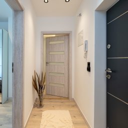 Patrycja Rozpondek NIERUCHOMOŚCI - Jasny korytarz z drewnianą podłogą i beżowym dywanem w kształcie rozgwiazd. Widoczne drzwi wewnętrzne z przeszkleniami i czarne drzwi wejściowe.