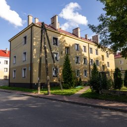 Patrycja Rozpondek NIERUCHOMOŚCI - Żółty blok z czerwonym dachem w słoneczny dzień. Widoczne drzewa i chodnik. Ujęcie z poziomu ulicy, z fragmentem samochodu w lewym dolnym rogu.