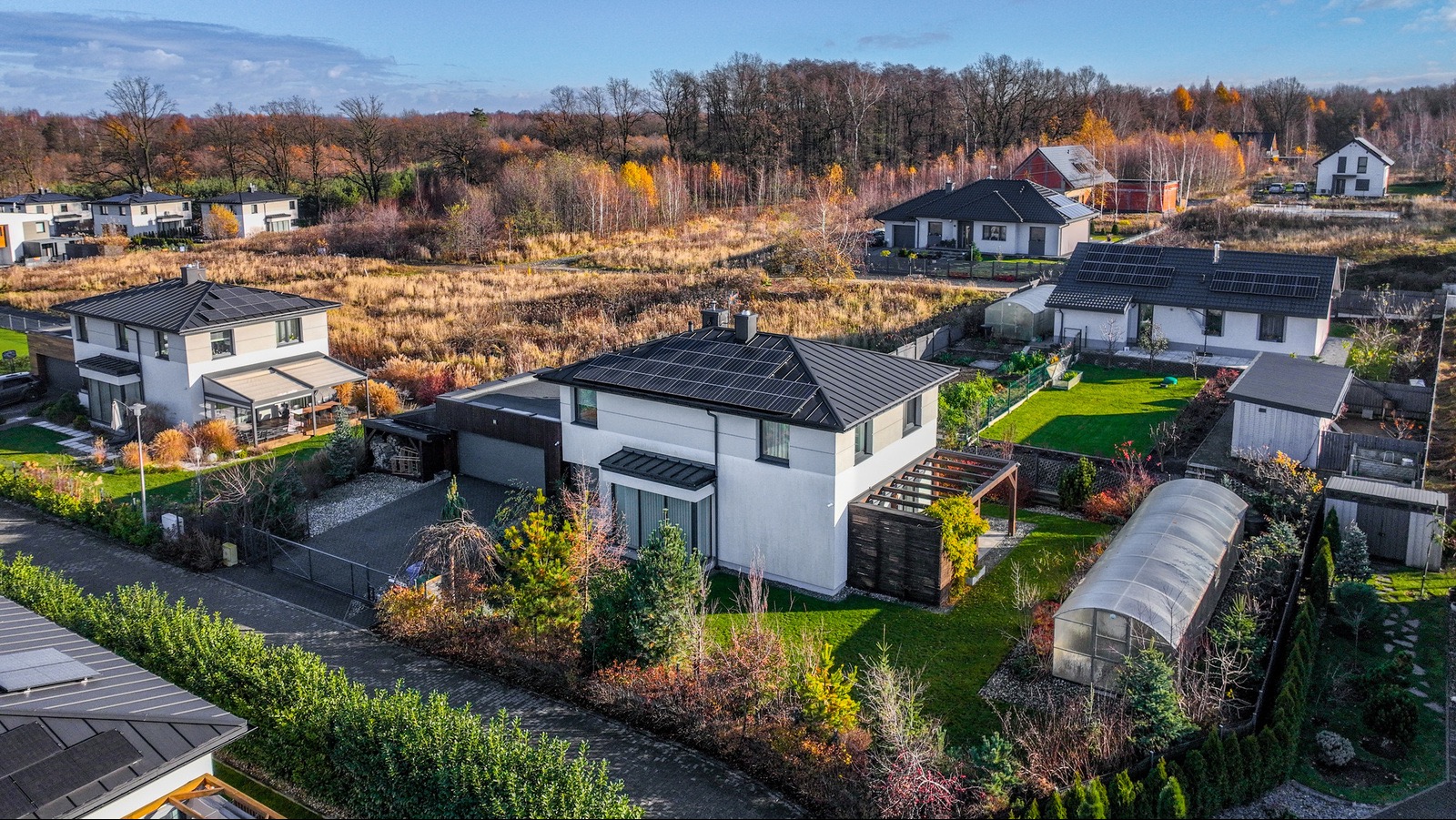 Nowoczesny dom jednorodzinny z panelami słonecznymi na dachu, widok z góry, jesienne otoczenie. W tle inne domy i drzewa.
