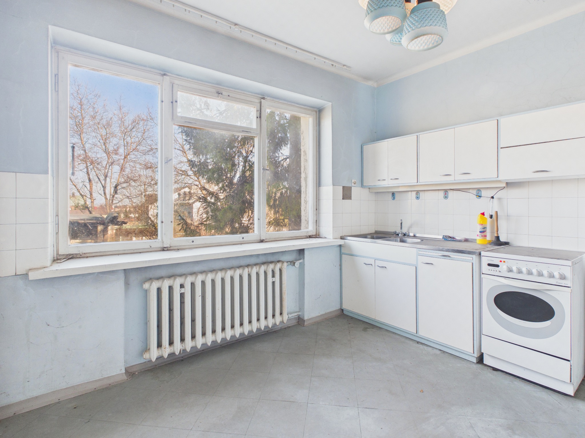 Jasna kuchnia w bloku z oknem na drzewa. Białe szafki, kuchenka i grzejnik. Błękitne ściany i szara podłoga. Widok z okna na drzewa bez liści.