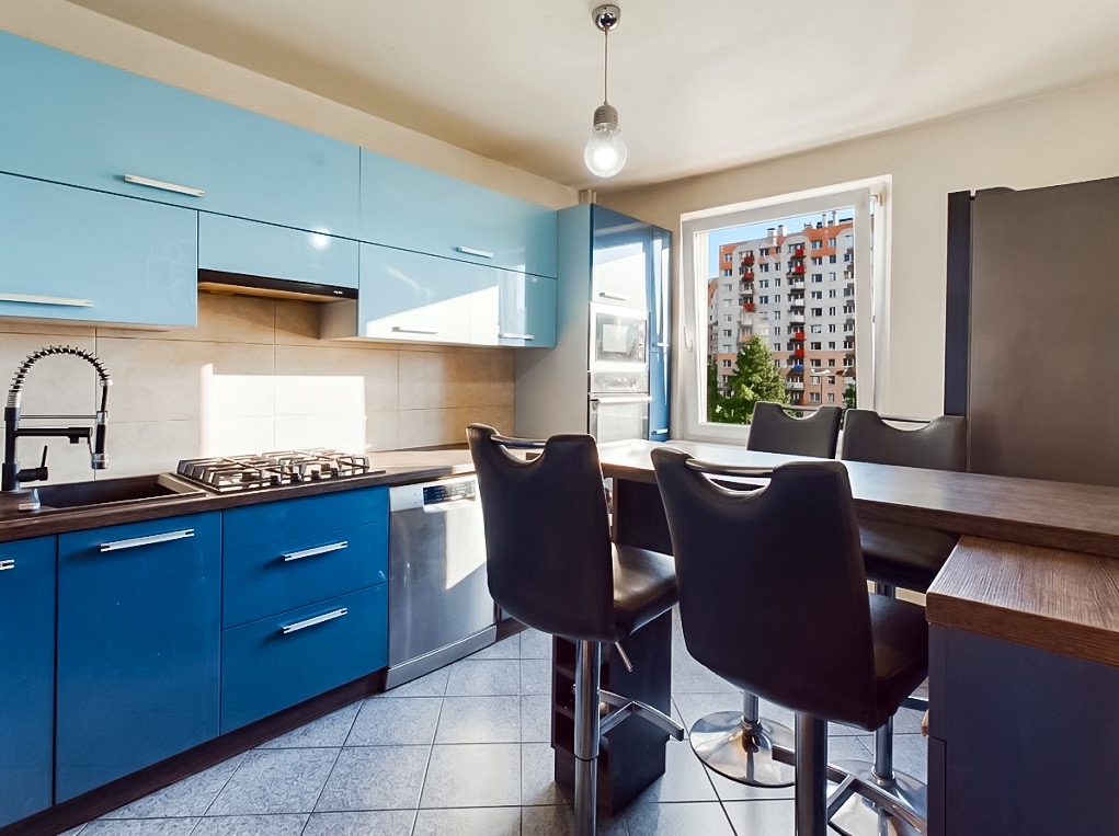 Błękitna kuchnia z wyspą i hokerami, widok na blok przez okno. Nowoczesne meble, płytki podłogowe. Lampa wisząca w stylu industrialnym. Jasne wnętrze.
