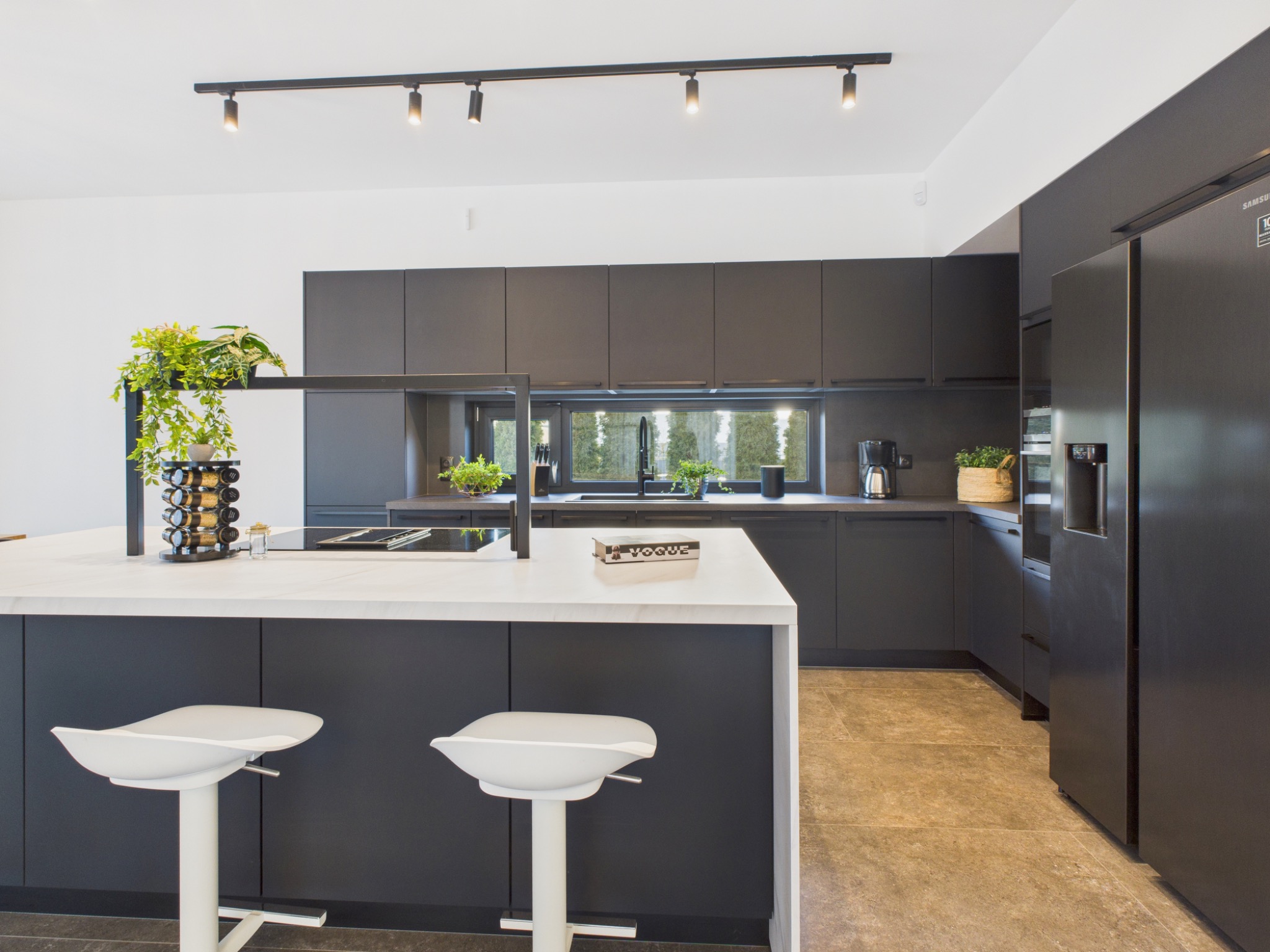 Nowoczesna kuchnia z wyspą i białymi hokerami. Czarne szafki, oświetlenie punktowe i marmurowa podłoga. Na wyspie książka Vogue i przyprawnik.