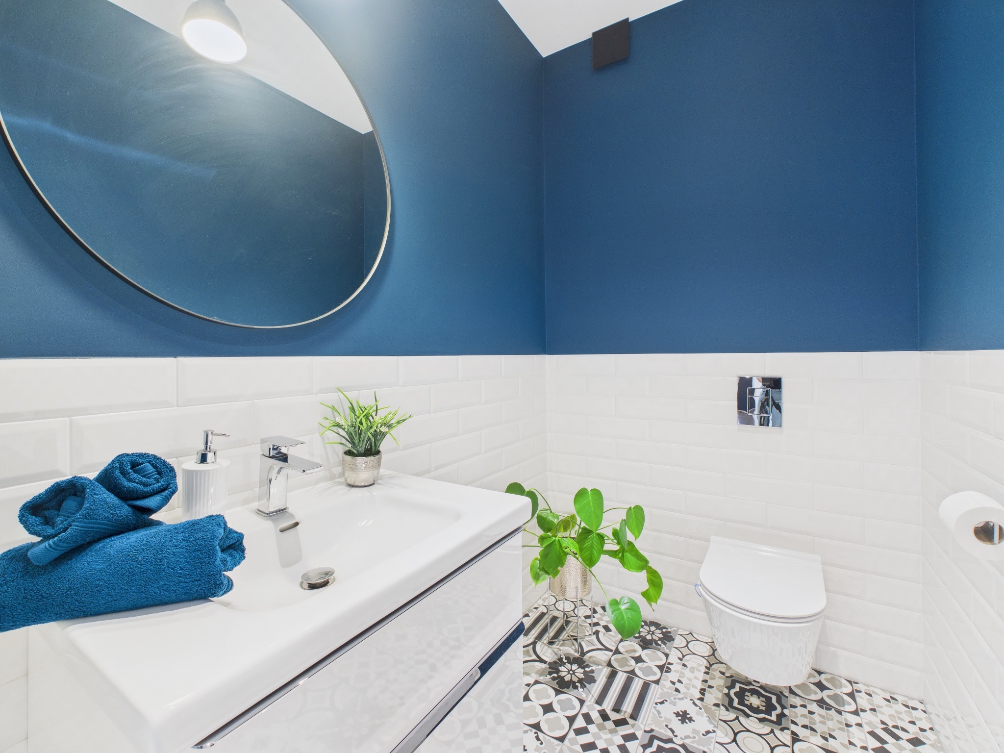 Nowoczesna łazienka z białą umywalką, okrągłym lustrem i akcentami w kolorze głębokiego błękitu. Dekoracyjne płytki podłogowe i rośliny dodają wnętrzu charakteru.