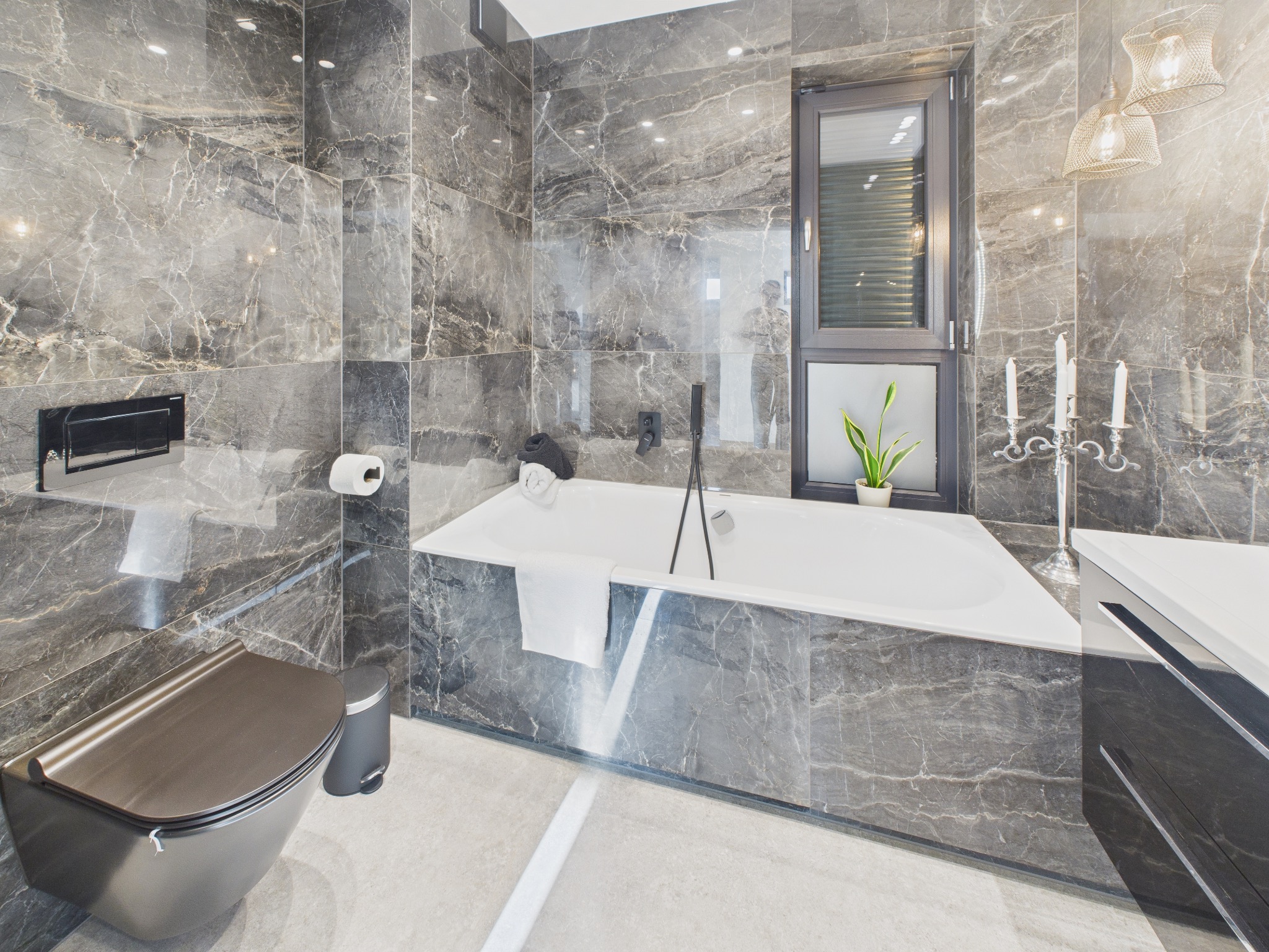 Elegancka łazienka z marmurowymi płytkami, wolnostojącą wanną, podwieszaną toaletą i oknem z roletami. Nowoczesny design z akcentami świec i roślin.