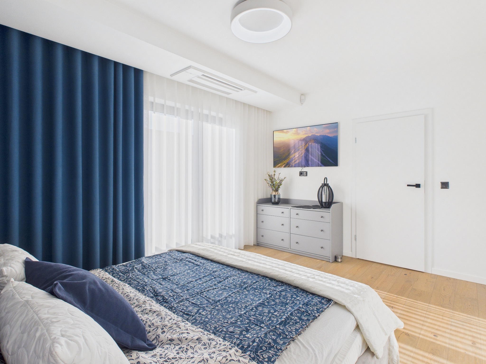 Jasna sypialnia z łóżkiem w niebieskiej pościeli, szarą komodą i telewizorem. Minimalistyczny design, drewniana podłoga, niebieskie zasłony i białe ściany.
