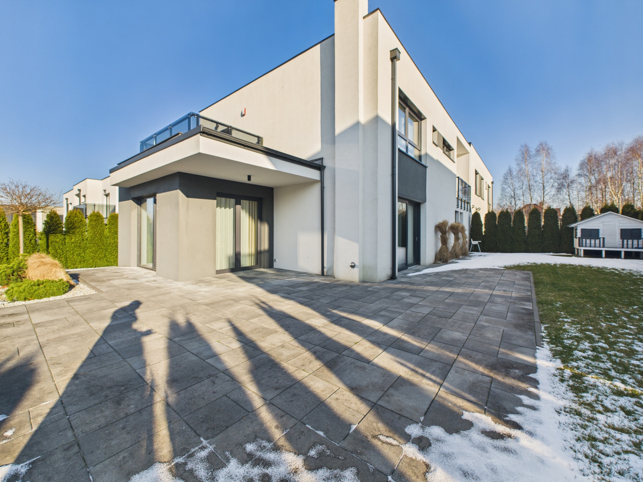 Nowoczesny dom z płaskim dachem i dużymi oknami, otoczony zielenią i częściowo pokryty śniegiem. Na pierwszym planie cień osoby na brukowanym podwórku.