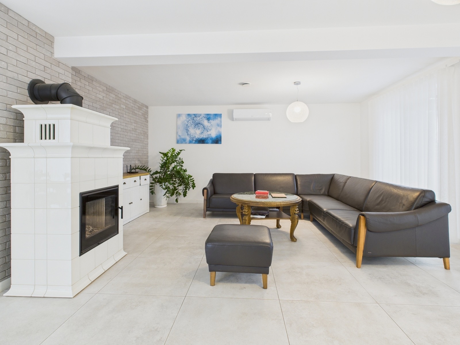 Jasne wnętrze salonu z białym piecem kaflowym, szarą cegłą na ścianie, skórzaną sofą narożną i okrągłym stolikiem kawowym. Minimalistyczny design z akcentami roślinnymi.
