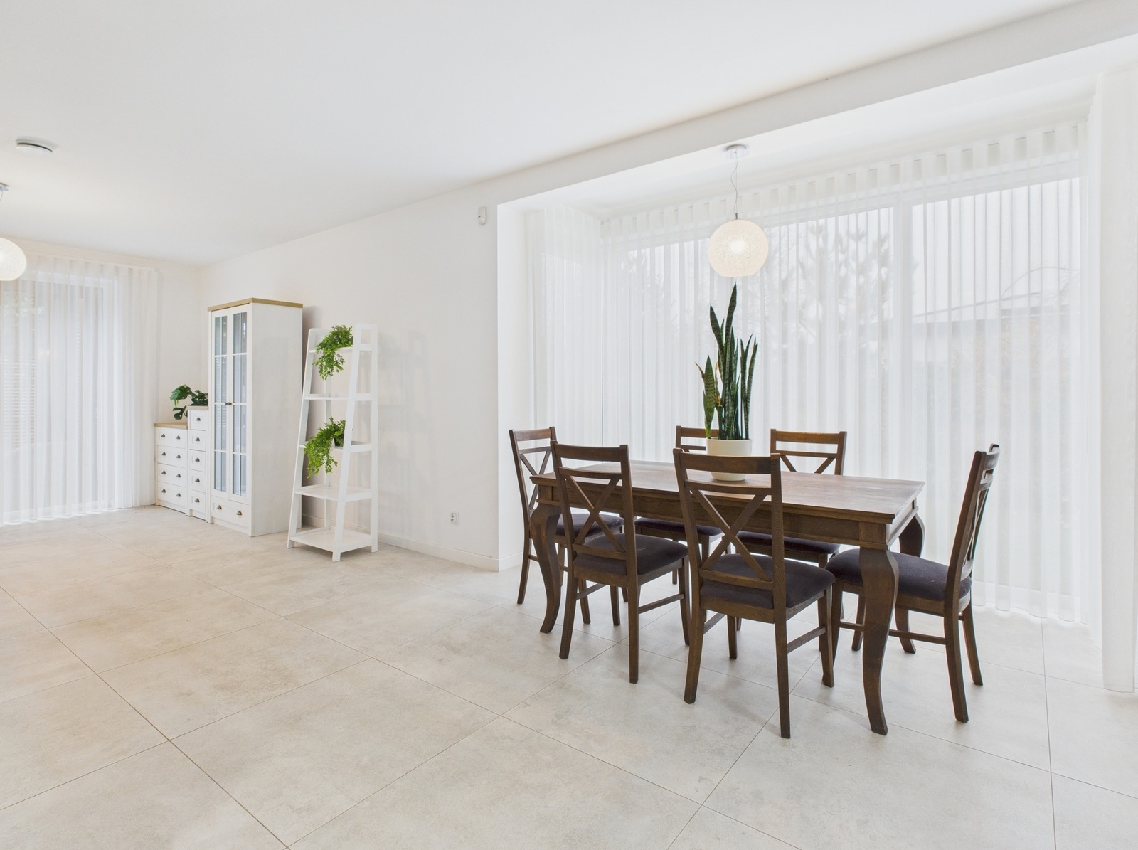 Jasne wnętrze jadalni z drewnianym stołem i krzesłami, oświetlone naturalnym światłem przez okno z białymi zasłonami. Minimalistyczny design z roślinami doniczkowymi.