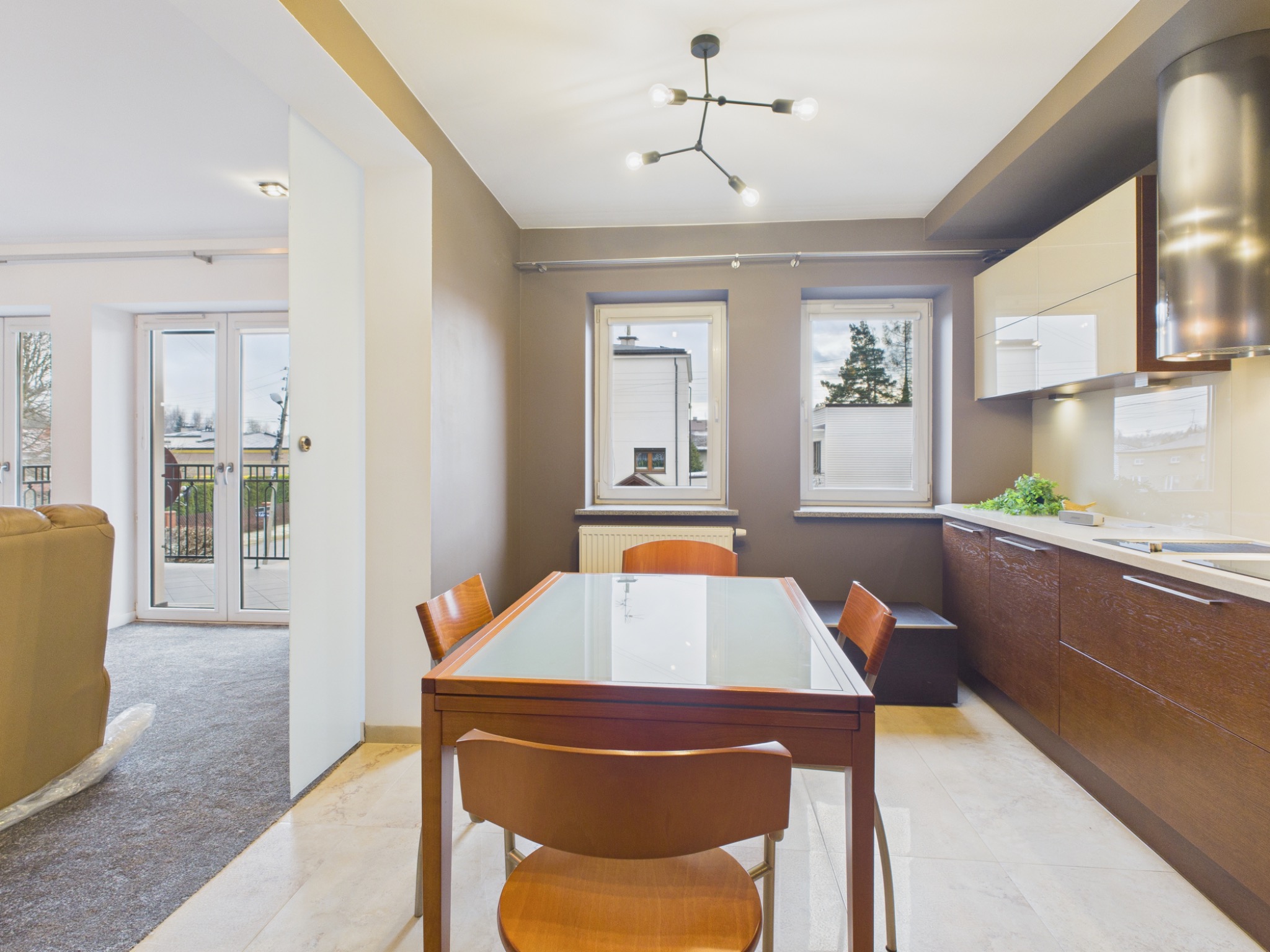 Jasna jadalnia z drewnianym stołem z szklanym blatem i krzesłami, widok na kuchnię z brązowymi szafkami i salon z balkonem. Nowoczesne oświetlenie sufitowe.