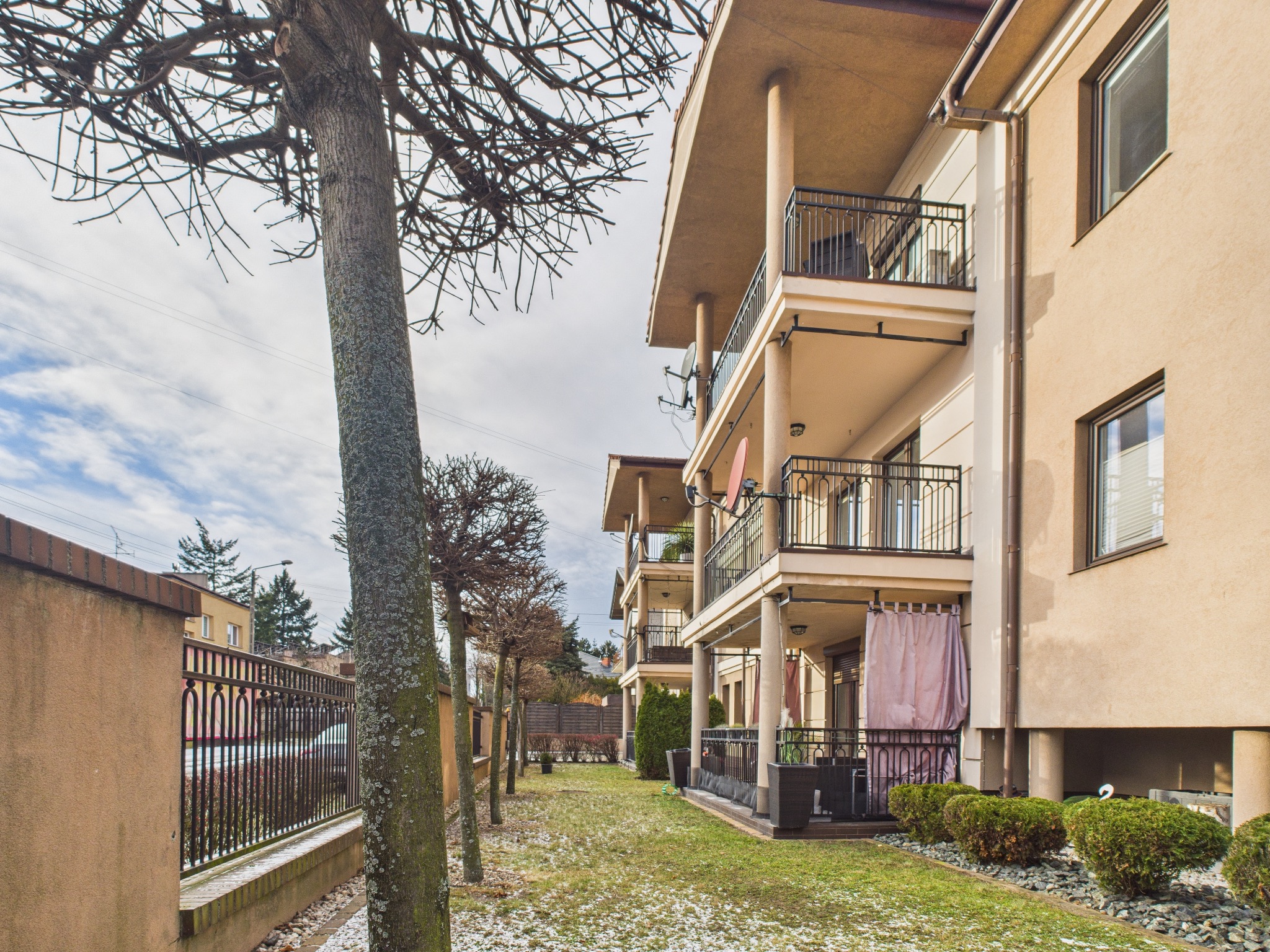Elewacja budynku wielorodzinnego z balkonami i zadbanym ogrodem. Widoczne drzewa bez liści i fragment ogrodzenia. Ujęcie z dołu, podkreślające wysokość budynku.