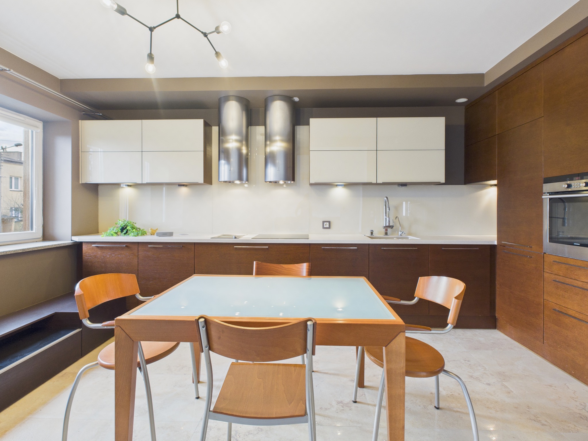 Nowoczesna kuchnia z drewnianymi szafkami, białymi frontami i szklanym stołem. Dwie srebrne tuby okapów nad płytą. Jasna podłoga z marmuru.