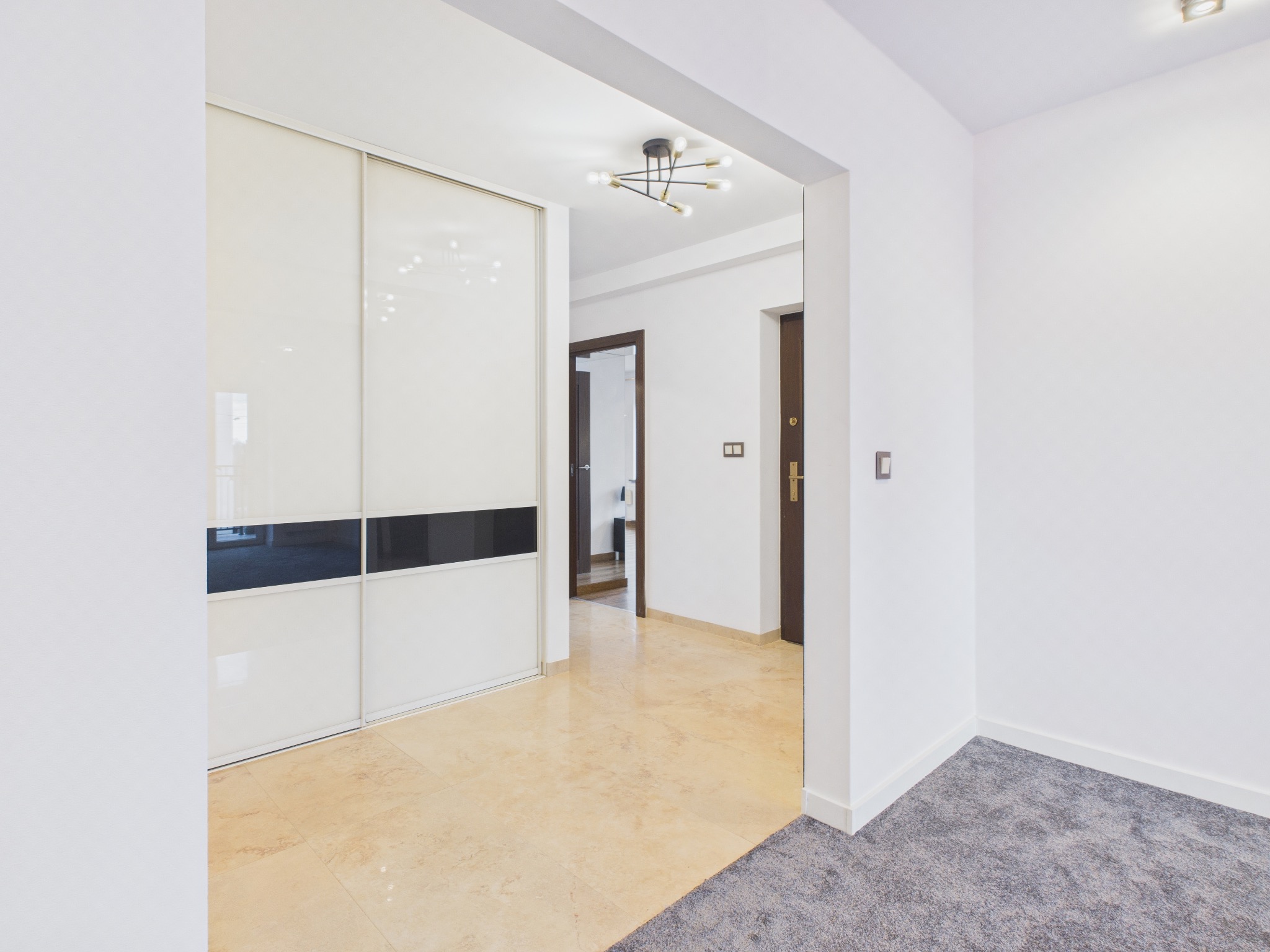 Jasne wnętrze mieszkania z beżową podłogą, białą szafą przesuwną z czarnymi akcentami i widokiem na dalsze pomieszczenia. Nowoczesna lampa sufitowa.
