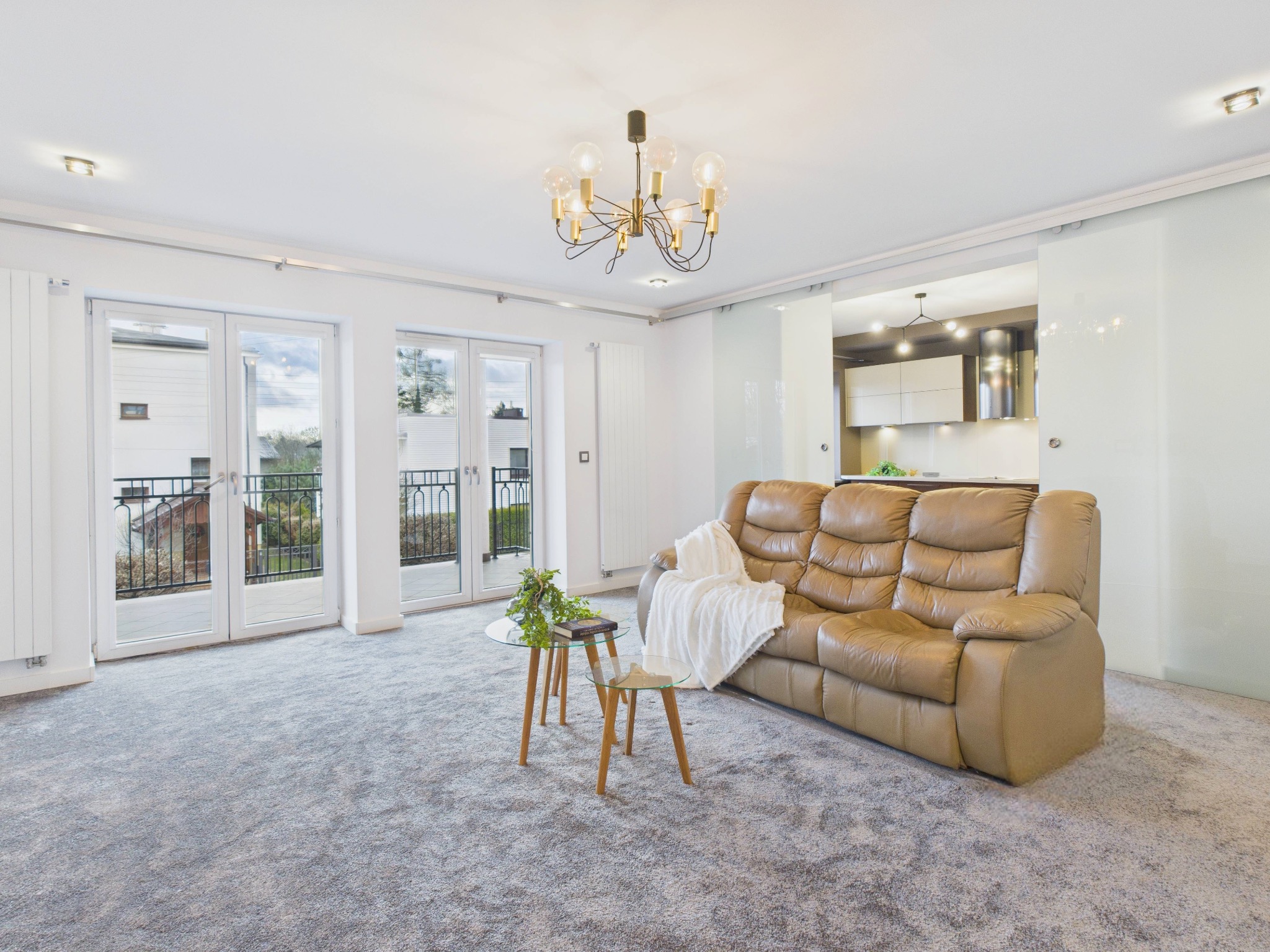 Jasne wnętrze salonu z beżową sofą, szarym dywanem i przeszklonymi drzwiami balkonowymi. Widoczny nowoczesny żyrandol i częściowo otwarta kuchnia.