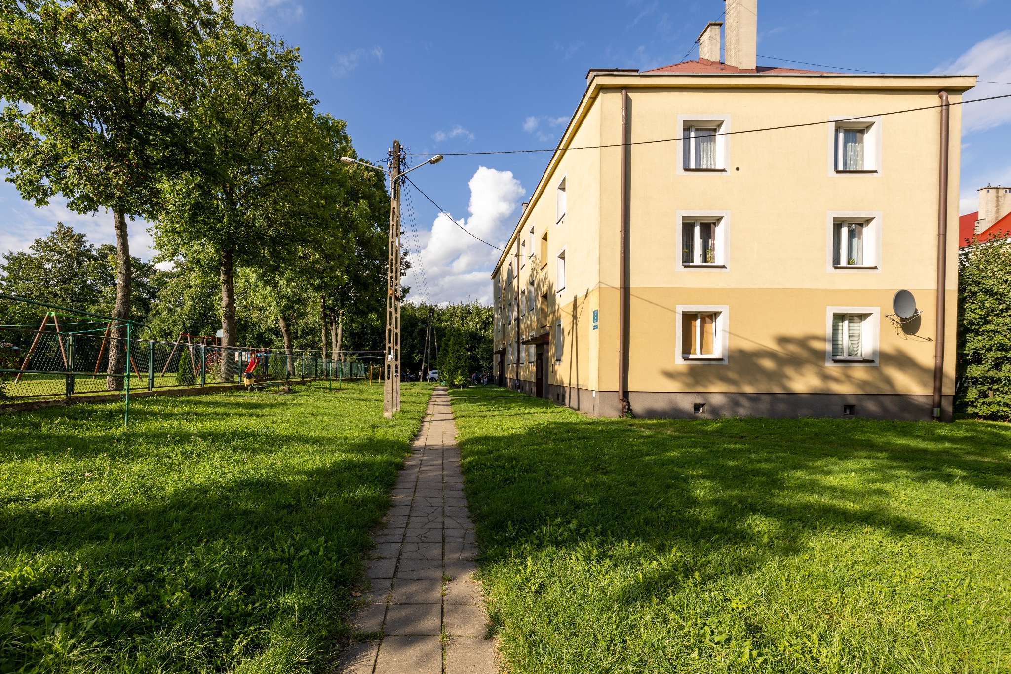 Żółty blok z anteną satelitarną, widziany z perspektywy ścieżki przez trawnik w słoneczny dzień. Widoczne drzewa i plac zabaw w tle.