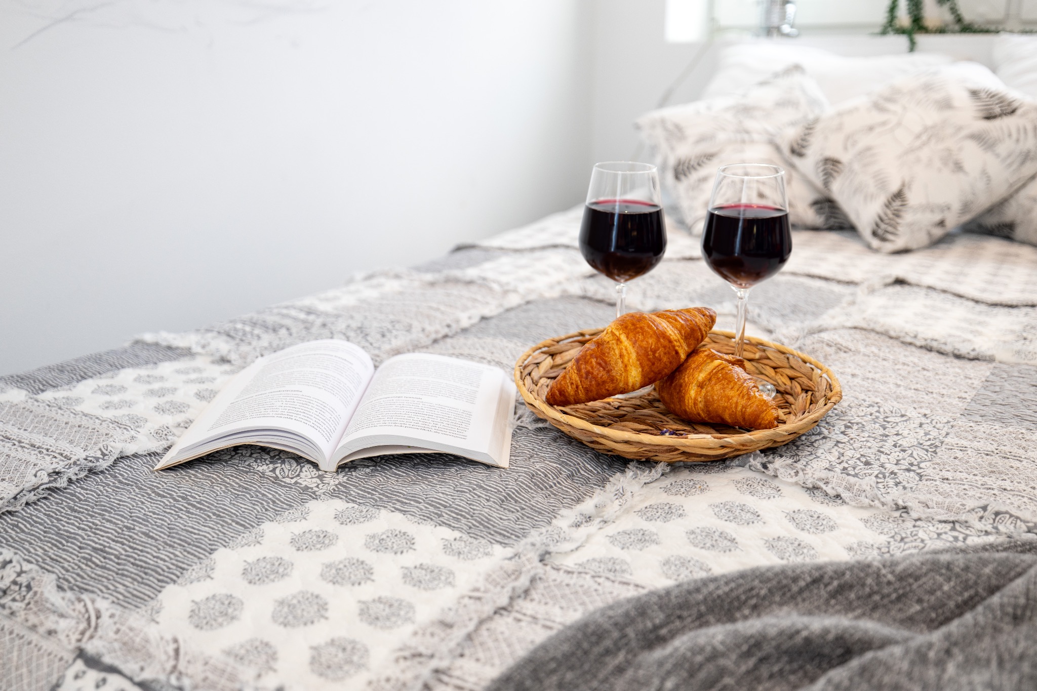 Śniadanie na łóżku: dwa rogaliki i kieliszki z czerwonym winem na plecionej tacy, obok otwarta książka na patchworkowej narzucie w odcieniach szarości i bieli.