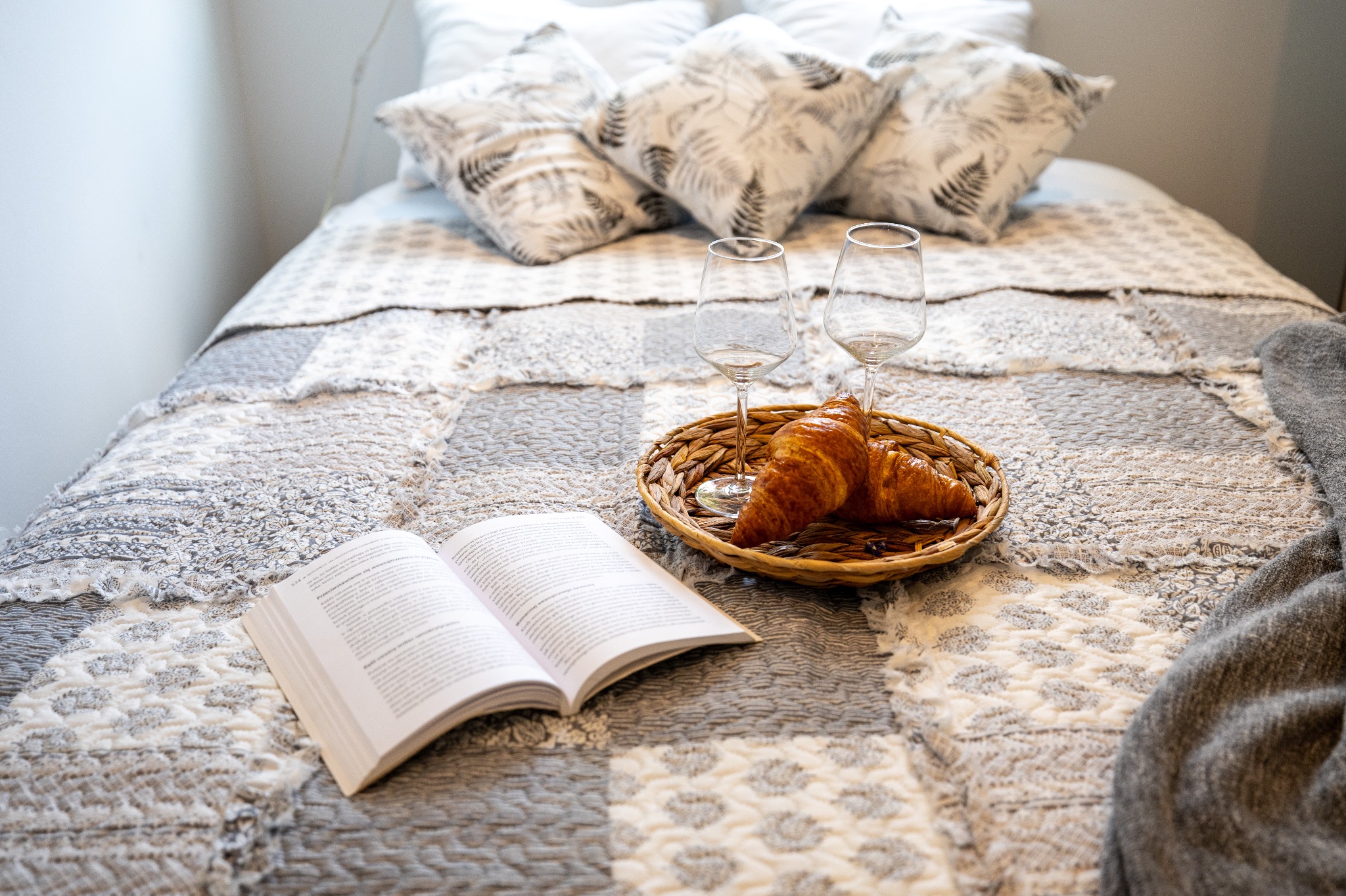 Śniadanie na łóżku: dwa rogaliki i kieliszki na tacy, otwarta książka na patchworkowej narzucie, poduszki w tle. Przytulna aranżacja w neutralnych barwach.