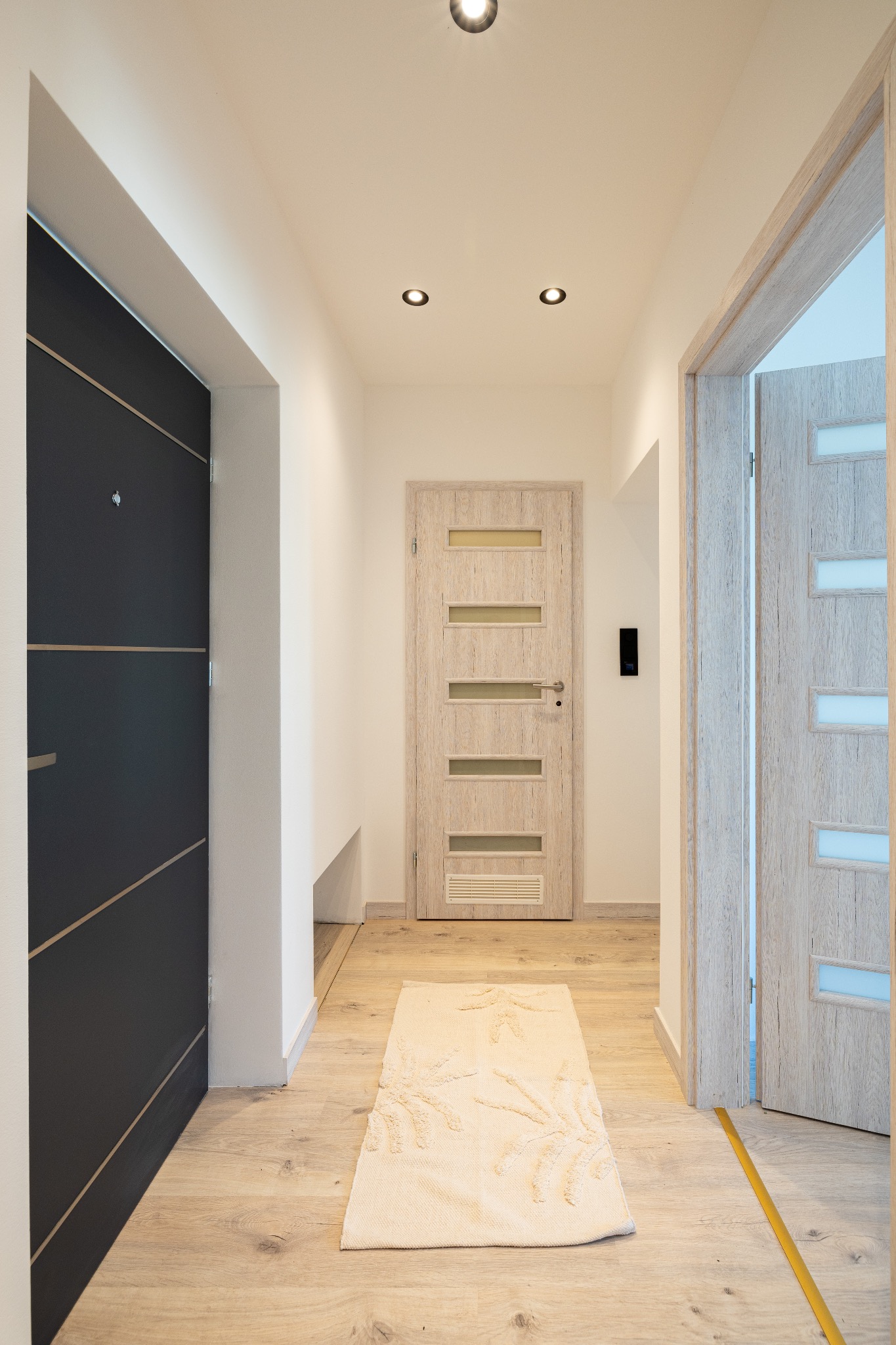 Nowoczesny korytarz z trzema drzwiami: ciemne wejściowe, jasne wewnętrzne z przeszkleniami i otwarte drzwi do pokoju. Beżowy dywanik na podłodze.