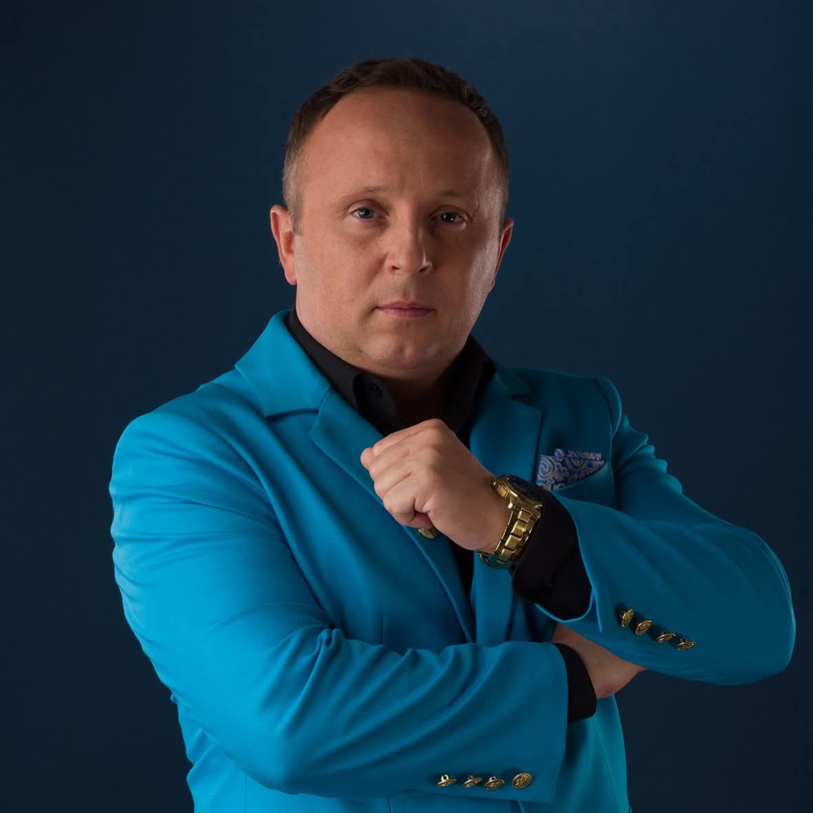 Mężczyzna w błękitnej marynarce i złotym zegarku na ciemnoniebieskim tle, z zaciśniętą pięścią. Portret biznesowy z nutą ekstrawagancji.