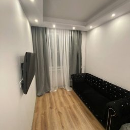MenArt Artem Menin - Wykończone wnętrze pokoju po remoncie: drewniana podłoga, czarna sofa z guzikami, telewizor na ścianie, okno z zasłonami. Nowoczesne oświetlenie sufitowe.