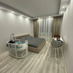 MenArt Artem Menin - Jasne wnętrze sypialni po remoncie: łóżko z szarym pledem, białe panele podłogowe, zasłony w odcieniach beżu i suszarka na pranie. Widoczne białe łóżeczko dziecięce.