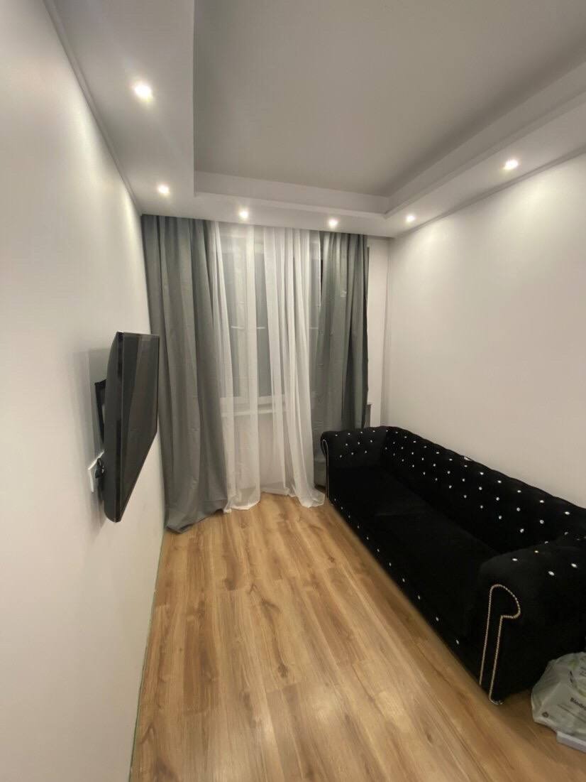 Wykończone wnętrze pokoju po remoncie: drewniana podłoga, czarna sofa z guzikami, telewizor na ścianie, okno z zasłonami. Nowoczesne oświetlenie sufitowe.