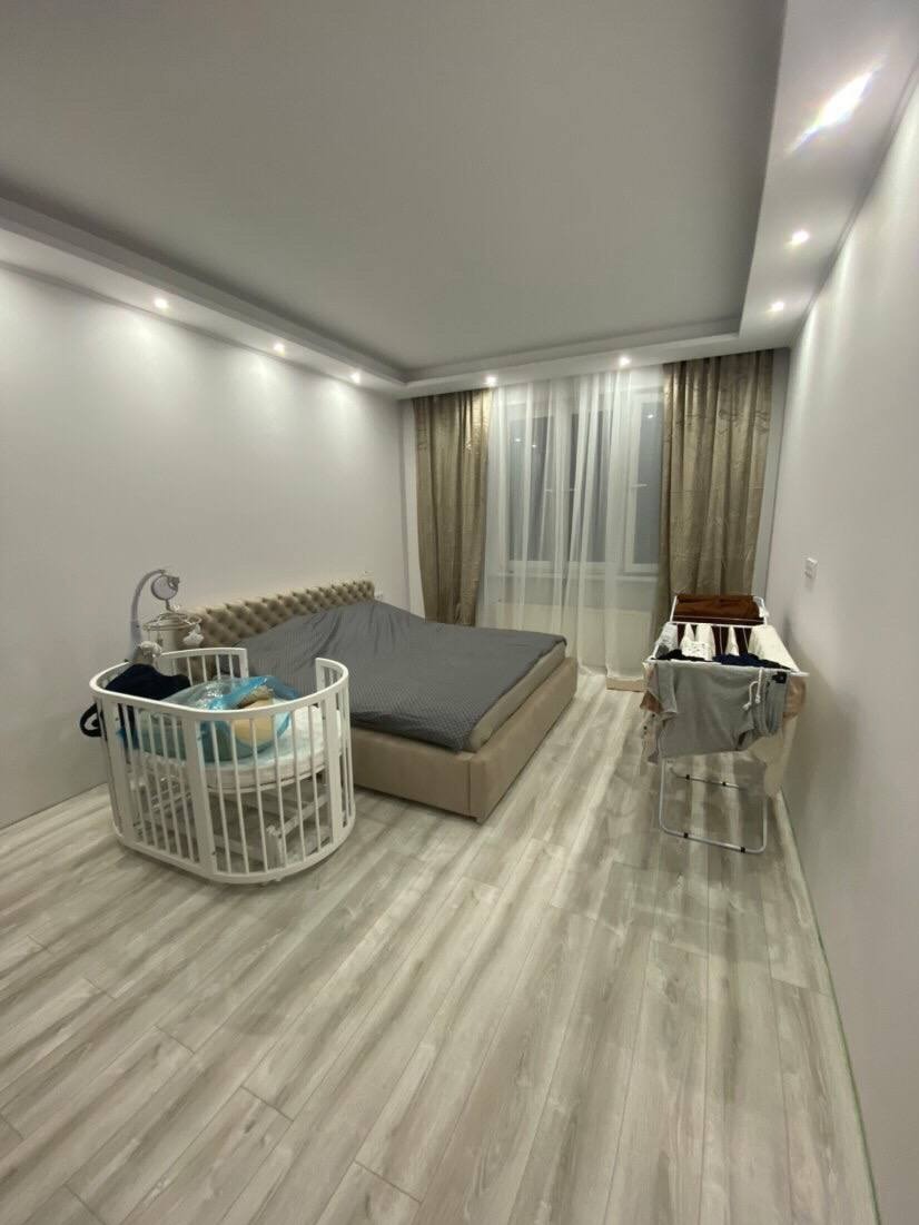 Jasne wnętrze sypialni po remoncie: łóżko z szarym pledem, białe panele podłogowe, zasłony w odcieniach beżu i suszarka na pranie. Widoczne białe łóżeczko dziecięce.