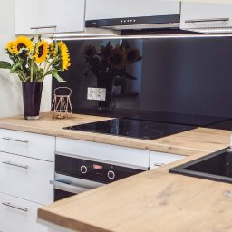 Duo Serwis - Nowoczesna kuchnia z białymi szafkami, drewnianym blatem i czarnym panelem szklanym. Na blacie wazon ze słonecznikami i dekoracje. Ujęcie z perspektywy.